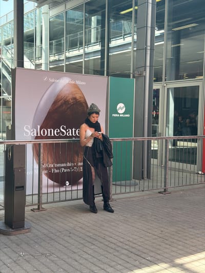 An outdoor, full-length shot shows a person posing confidently in front of a large promotional poster at the Fiera Milano exhibition center in Rho, Italy. The setting features a modern building with a glass and metal facade reflecting the bright, sunny sky. The ground is paved with grey interlocking bricks, and metal barriers separate the paved area from the poster and building.
The subject is dressed in a stylish black and white ensemble, including a white polka-dot top, a black jacket or coat, black trousers, and black boots. They wear a distinctive striped black and white head covering and accessorize with a necklace and rings. The person is leaning slightly against a metal barrier, holding what appears to be a phone in one hand, and looking towards the camera.
The large poster prominently displays "Salone del Mobile.Milano" and "SaloneSatellite," indicating a major design or furniture fair. Another green sign for "FIERA MILANO" is visible behind the barriers. Partial text on the poster also reads "Skied Craftsman ship + Inn" and "era Mino - Fho (Pavs 5-7) | 2." The bright sunlight suggests it is daytime, with clear shadows cast on the pavement. The overall scene captures a moment at a large-scale exhibition or event.