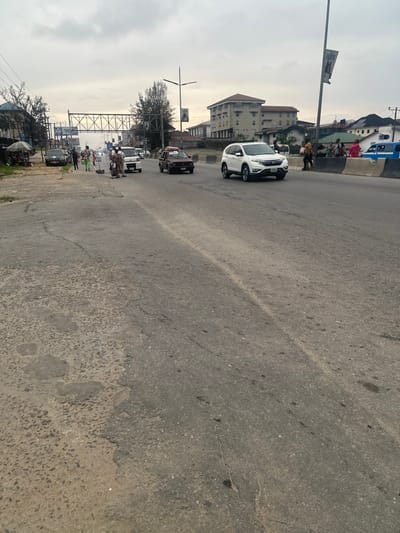 A street view shows a wide road with a white SUV and other vehicles further down. On the sides, people walk and structures such as buildings and utility poles are visible. The road surface is cracked and patched, suggesting wear and tear. The sky is overcast, casting a soft, diffused light over the scene.