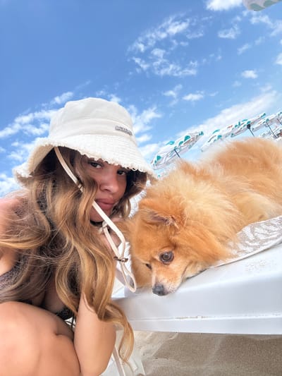 A woman and a fluffy dog are on a beach, possibly at Primorsko, Bulgaria. The woman is wearing a white bucket hat and a sparkly top. Her long blonde hair is visible, framing her face and cascading over her shoulder. She is looking towards the camera with a relaxed expression.
The dog, a Pomeranian with golden fur, is lying down next to her. Its head is turned away from the camera, but its alert eye is visible. They appear to be relaxing on a lounge chair. In the background, a bright blue sky with scattered white clouds is visible, suggesting it's daytime and the weather is pleasant. Some beach umbrellas with a decorative pattern are also partially visible. The sand of the beach is seen on the right side of the image. The overall mood is one of leisure and enjoyment on a sunny day at the beach.