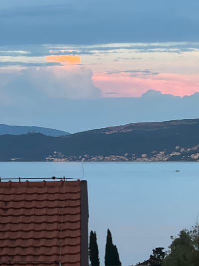 The image captures a serene twilight scene over the Bay of Kotor, specifically looking towards the mountainous coastline near Baošići, Montenegro. In the foreground, the terracotta tiled roof of a building is visible, hinting at a residential area. The middle ground is dominated by the calm, pale blue water of the bay, with a distant, small boat barely visible on the surface. Across the bay, a dark, heavily wooded mountain range forms the backdrop. The slopes of the mountains are dotted with small white structures, suggesting a coastal town or villages nestled along the shore. The sky above is a gradient of soft blues and pale pinks, with clouds tinged with orange and yellow from the setting sun or moon. A notable feature in the sky is a bright, horizontal band of light, possibly a lenticular cloud illuminated by the sun or moon. The overall atmosphere is peaceful and tranquil, characteristic of an early evening in a coastal Mediterranean setting.