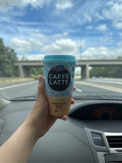 A hand holds a light blue and beige paper cup of coffee latte while inside a car. The cup has text that reads "CAFFÈ LATTE SKINNY". The car's dashboard is visible in the foreground, with the steering wheel partially obscured. Outside the car, a highway overpass is visible above a tree line, with a cloudy blue sky in the background. The scene is outdoors on a sunny day. The location is not identifiable as Leeds, United Kingdom, or any other specific city.