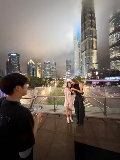 The image captures a nighttime scene in Pudong, China, with a panoramic view of the city's iconic skyscrapers illuminated against a hazy sky. In the foreground, two young women are posing for a photograph. One woman, wearing a black sleeveless top and dark pants, is embracing the other, who is in a light-colored dress. They appear to be sharing a tender moment, possibly friends or a couple, with smiles on their faces.
The background is dominated by the towering, modern architecture of Pudong's financial district. Buildings like the Jin Mao Tower and Shanghai World Financial Center are visible, their facades shimmering with countless lights, reflecting the urban vibrancy of the city. The time of day is clearly night, with artificial lighting being the primary source of illumination. The weather appears slightly overcast or misty, contributing to the diffused glow of the city lights.
A person's head and shoulder are visible in the lower right corner of the image, suggesting the photographer or someone else in the group, looking towards the city skyline. They are holding a smartphone, presumably to capture the moment or the view. The setting appears to be an elevated vantage point, perhaps a rooftop bar or a viewing deck, offering an expansive perspective of the cityscape. The overall mood is one of modern urban exploration and shared experience, with a touch of intimacy between the two women. No discernible text is visible in the image.