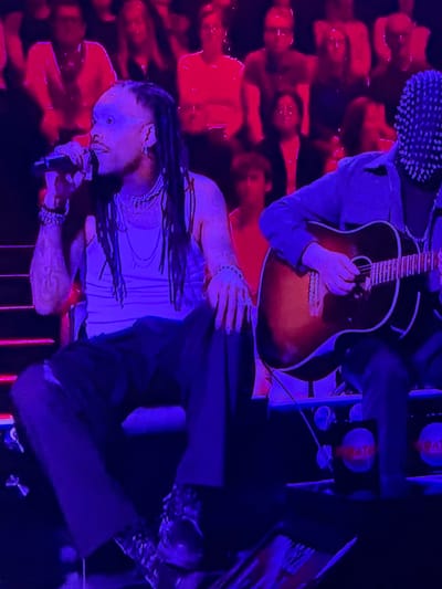 A music performance is in progress in Marche-en-Famenne, Belgium. A person with dreadlocks is singing into a microphone, wearing a white shirt and standing in front of a red illuminated crowd. Above them, a guitarist plays an acoustic guitar. To the right of the vocalist, stands a table stacked with what appears to be merchandise. A person wearing a studded cap is visible in the upper left corner. The environment is bathed in red and blue stage lighting.