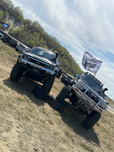 The image shows a gathering of off-road vehicles, primarily Toyota Hilux models, in a dirt/grassy area. License plates indicate the location as Russia. A "HILUX SURF CLUB" flag is attached to one vehicle. The vehicles are parked in a line, with a wooded area and a rocky outcrop visible in the background. The location could be near Sadovoe, Russia, but there is no definitive information to determine the exact location. The image does not contain information about any people, actions, or interactions.