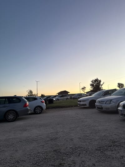 The image shows a line of parked cars in a lot. The location is likely Lukovit, Bulgaria. The sky is a gradient of blue to orange, suggesting dawn or dusk. There is a wind turbine visible in the background. The ground is gravel. No people are visible.