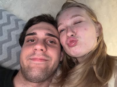 A close-up, overhead selfie captures a young Caucasian couple, a male on the left and a female on the right, in an indoor setting. The male has dark hair and a light complexion, with visible stubble on his face. He is looking directly at the camera. The female has blonde hair and a fair complexion, with some minor blemishes on her skin. She is winking and puckering her lips towards the camera. They are positioned very close together, with their heads nearly touching. The background shows a textured wall and a grey, chevron-patterned blanket or cushion on the left. The lighting appears to be warm and ambient. There are no obvious indicators of the specific location within Rossiyskiy, Russia, beyond the general indoor environment.