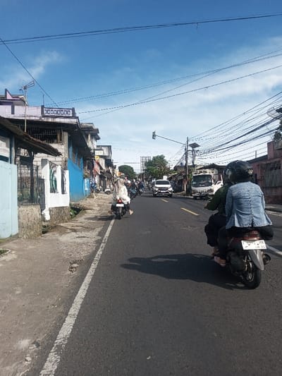The image displays a street scene in the city of Phonengam, Laos, observed on a clear day with a blue sky and sparse white clouds. The asphalt road, appearing to be a single direction of travel, recedes into the distance. In the immediate foreground on the right, a dark-colored scooter is ridden by an individual in a dark green jacket and black helmet, carrying a passenger in a light blue denim-style jacket and black helmet. The scooter's license plate is "F 6392 MK AN 36". To their left, another individual, dressed in a white top and light pants, operates a light-colored scooter. Further along the road, several additional scooters, a white SUV, and a white truck are visible, all moving away from the viewer.

On the left side of the road, a concrete sidewalk abuts a series of multi-story buildings, some painted in faded light blue and white, featuring barred windows and metal awnings. An antenna is present on one rooftop. Overhead, numerous utility wires crisscross the sky, connected to poles positioned on both sides of the street. A single traffic light hangs suspended above the road in the mid-ground. Shadows from vehicles and individuals are cast onto the road surface.