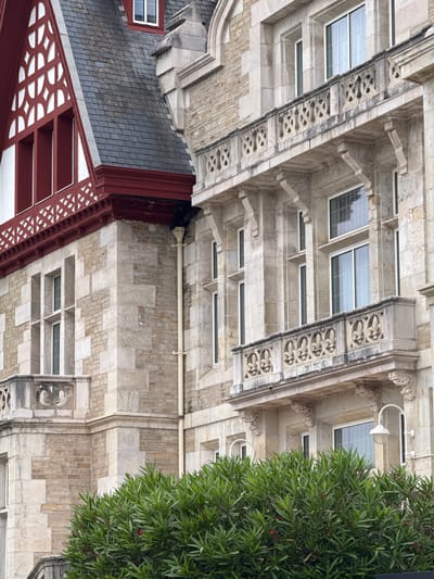 The image captures a close-up view of a section of the Palacio de la Magdalena in Santander, Spain. The architecture features a mix of stone and red-painted elements, with a prominent slate roof on the left side. Elaborate stonework adorns the balconies, which have decorative railings. Several windows, some with white frames and gridded panes, are visible on the stone facade. A lush green bush with small pink flowers occupies the foreground, partially obscuring the lower portion of the building. The lighting suggests an overcast or diffused daylight condition. No people are present in the scene.