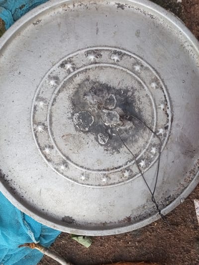 The image displays a close-up, overhead view of a weathered, circular metal object, likely a pot or a large pan, resting on the ground. The object is silver-toned and shows signs of age and use, with dark, stained areas concentrated in the center and along the rim. A decorative pattern of embossed stars is visible on concentric rings towards the center of the object. Two dark wires are loosely tied together and rest on the surface of the metal, extending towards the right side of the frame. The background consists of a patch of rough, sandy ground, a portion of a blue mesh material in the lower left, and some dry leaves or debris. The overall impression is one of an outdoor setting, possibly in a less maintained area. There is no identifiable specific location within Zinna, Nigeria provided by the image itself.