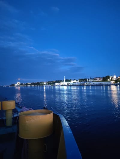 The image depicts a body of water at dusk or early night, with the lights of a city illuminated along the distant shore. In the foreground, parts of a vessel are visible, including yellow cylindrical objects and a blue railing. The water reflects the lights from the city, creating shimmering patterns. The sky is a deep blue with some scattered clouds. The city skyline features buildings and a prominent white structure that appears to be a church or a minaret. The overall impression is one of a calm evening scene on a waterway adjacent to a populated area. The location is identified as Shebunino, Russia.