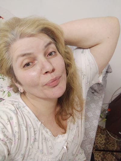 A woman with blonde hair and light-colored eyes lies in bed, resting her head on her arm. She is wearing a white nightgown with a floral pattern and lace trim. The background shows a white wall and a patterned blanket. The image does not provide any information about the location within Al Mas'oudi, United Arab Emirates.