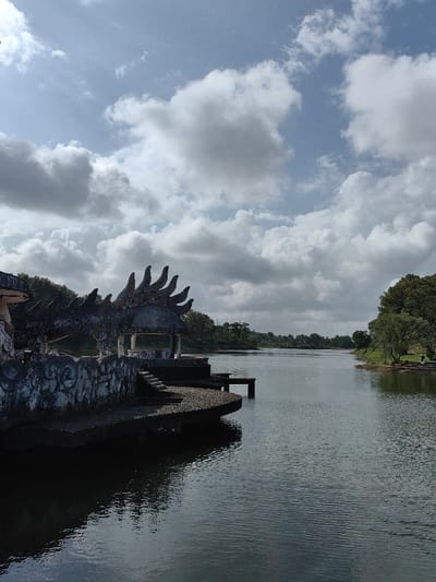 The image displays an exterior scene in Kim Long Ward, Vietnam, featuring a body of water with structures along its left bank under a partly cloudy sky. The central and lower portions of the frame are occupied by a dark, placid body of water, likely a lake or wide river, exhibiting surface ripples and reflections of the sky and surrounding elements.
On the left side, a weathered concrete structure extends from the bank into the water. This structure is sculpted with features resembling a dragon, including a series of prominent, fin-like crests along its upper edge. Integrated into this construction is a circular, domed gazebo with supporting pillars. Both the dragon structure and the gazebo exhibit significant weathering, discoloration, and graffiti markings on their surfaces. A set of concrete steps leads down from the main platform of the structure towards the water, and a smaller, rectangular concrete pier projects further into the water from this platform.
The right bank of the water is densely covered with green trees and foliage, extending into the mid-ground. Additional trees line the horizon in the background on both sides of the water. The sky above is a light blue, extensively covered by numerous large, white and grey cumulus clouds. No individuals, animals, or explicit actions are depicted in the scene.