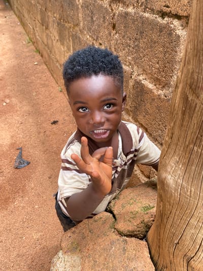 The image, taken in Kuru Dungus, Nigeria, captures a cheerful young boy in an outdoor, natural setting. The boy, appearing to be of African descent, is the central subject, viewed from an overhead perspective looking up at the camera. He has short, dark, curly hair and a bright smile, revealing his teeth, with wide, engaged eyes. He is wearing a light-colored t-shirt with brown stripes and dark-colored shorts or trousers. His right hand is raised towards the camera, fingers slightly splayed, in what appears to be a friendly gesture or greeting.
The background features large, rough-textured reddish-brown rocks, some covered with patches of green moss, suggesting a natural and somewhat rustic environment. A weathered wooden log or beam with prominent grain runs horizontally across the upper part of the frame, positioned behind and above the boy. The ground is reddish-brown dirt or sand. A small, dark, crumpled piece of debris, possibly a leaf, is visible on the ground near the boy's lower right. The scene is illuminated by bright natural daylight, indicating daytime. There is no visible text in the image.