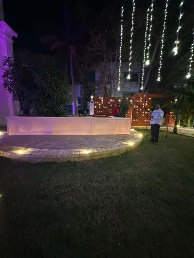 The image depicts a nighttime outdoor scene, consistent with an event or celebration setting. The specific location within Vadodara, India, is not discernible from the visual information provided.
In the foreground, a dark green grass lawn extends across the lower portion of the frame. A semi-circular raised platform, paved with light-colored stone tiles and bordered by brick, is positioned centrally. Small, embedded warm-toned spotlights illuminate the edge of this platform.
In the midground, on the raised platform, a long, rectangular structure, appearing to be a stage or table, is covered with a translucent light-colored fabric, illuminated from within or beneath with a soft glow. Behind this structure, slightly to the right, a male individual in a red shirt is partially visible, seated or crouching behind a small green plant. To his right, a decorative reddish-brown wall or partition features multiple square and rectangular cutouts through which warm light emanates. Standing on the grass to the right of the platform, another male individual wears a light-colored, patterned shirt and dark trousers, facing towards the left. He holds a dark, thin object vertically in front of him.
The background features numerous strings of small, warm white fairy lights hanging vertically, creating a canopy effect. Trees and dense foliage are visible behind the decorative wall and stage area, with some foliage also adorned with small lights. A portion of a light-colored building with architectural details, possibly illuminated with a purple uplight, is visible on the far left. The sky is dark, indicating night.