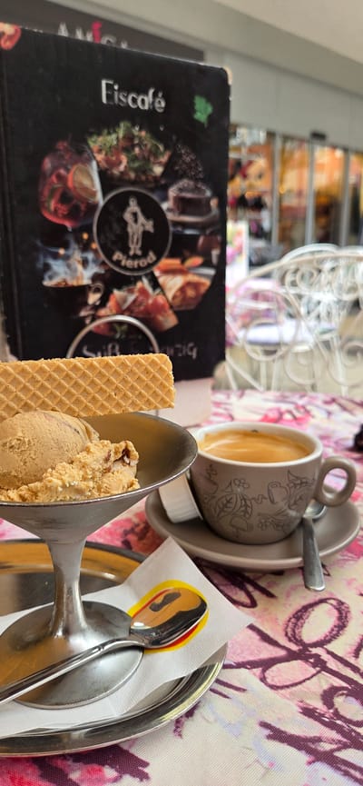 The image captures a close-up view of an ice cream sundae and a cup of espresso on a table, set in Kehl, Germany, with the facade of "Eiscafé Pierod" visible in the background, slightly out of focus. The ice cream is a light brown color, served in a metallic goblet with a wafer cookie perched on top. Next to it, a small, dark grey cup with a floral pattern holds a frothy espresso, placed on a matching saucer with a spoon. A white napkin with a yellow and red circular design is positioned beneath the spoon. The table is covered with a pink and white patterned tablecloth. The background shows the entrance to the cafe, with large glass doors and windows, hinting at an indoor seating area. The overall ambiance suggests a pleasant cafe setting in the city of Kehl. There are no people visible in the image.