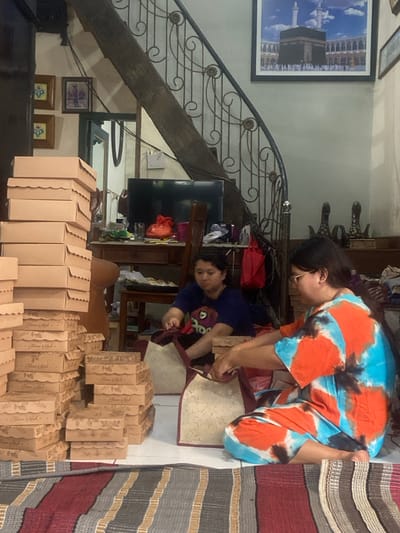 An indoor scene in Mojokerto, Indonesia, depicts two women engaged in packing activities within what appears to be a residential living area. In the foreground, a woman wearing a blue and orange tie-dye top and glasses is seated on a light-colored tiled floor, actively placing items into a patterned, maroon-trimmed tote bag. Behind her, another woman in a dark blue t-shirt is similarly engaged with a second identical tote bag.
To their left, a substantial quantity of light brown cardboard boxes with scalloped-edge lids, some marked "Finess Kesh," are stacked. A tall stack extends vertically on the far left, while multiple smaller stacks are arranged horizontally on the floor. A striped rug in shades of red, grey, and brown covers the immediate foreground.
The background reveals a domestic environment. A wooden staircase with an ornate dark metal railing ascends from the right side of the frame. Above the staircase, a large framed picture of the Kaaba in Mecca is mounted on a light-colored wall. Below the staircase, a dark wooden console or desk holds a flat-screen television, a red bag, and various miscellaneous items. Further to the left, two smaller framed pictures with blue and white designs are visible on the wall. A dark wooden chair is positioned behind the second woman, and a doorway is partially visible in the deep background. Decorative, dark-colored stylized figures are present on a surface to the right of the desk.