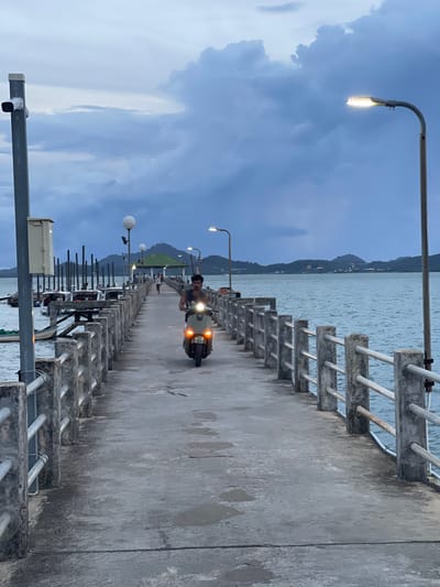 The image depicts a person riding a scooter down a pier. The scene appears to be a waterfront location, likely a harbor or a coastal area, given the presence of the sea and distant hills. The time of day is likely dusk or early evening, as indicated by the soft lighting from streetlights on the pier and the somewhat overcast sky. The weather appears to be cloudy. The pier itself is made of concrete and has a railing with metal bars. There are several streetlights along the pier, illuminating the path. In the background, hills covered in vegetation can be seen rising from the water, and there are some buildings visible along the coastline. The scooter has its headlights on, casting a glow on the wet-looking pavement. The person on the scooter is looking forward and appears to be alone. The overall atmosphere is serene and tranquil. No visible text is present in the image. The location context of Pa Khlok, Thailand, suggests a tropical coastal environment.