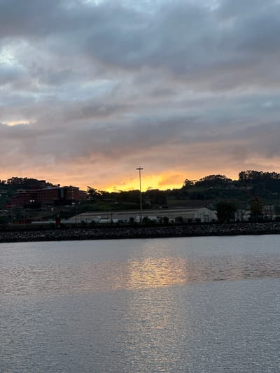 The image depicts a waterfront scene at dusk or dawn, likely in the city of Gijón, Spain, with the sun setting or rising behind a hill covered in trees and buildings. In the foreground, a calm body of water reflects the muted colors of the sky, which is dominated by gray and orange clouds. Along the horizon, a stone embankment separates the water from the land. Beyond the embankment, a large, multi-story brick building is visible on the side of the hill, with other industrial or commercial structures closer to the water. A tall light pole stands prominently on the right side of the image, and a crane can be seen further to the right, suggesting an industrial or port area. The overall atmosphere is tranquil and slightly overcast, with the warm hues of the sky providing a touch of color against the muted tones of the landscape. There are no people visible in the image.