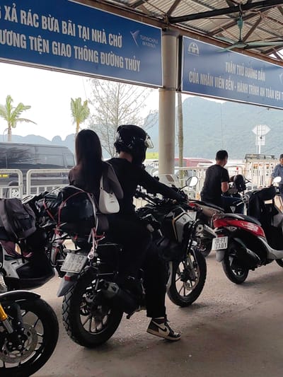 The image shows a scene at the Tuan Chau Marina in the city of Hải Phòng, Vietnam. Several motorcycles are parked under a covered area with signage in Vietnamese. One sign reads "TUANCHAU MARINA" and includes information about proper disposal of trash and safety instructions. A couple is on one of the motorcycles, with the rider wearing a helmet and the passenger carrying a white purse. The motorcycle is loaded with luggage. Other people are visible nearby. The background features mountains and a cloudy sky.