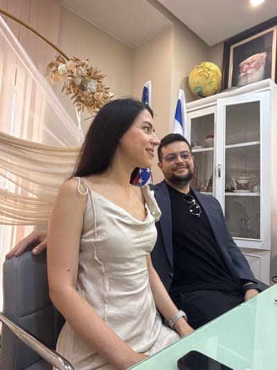 An indoor photograph, rotated 90 degrees clockwise, captures a man and a woman in what appears to be an office or decorated room in Rishon LeZion, Israel.
The man, with a beard, short dark hair, and eyeglasses, is positioned slightly behind the woman and looks directly at the camera with a gentle smile. He wears a black t-shirt underneath a dark blazer. A pair of silver-rimmed sunglasses is tucked into his t-shirt collar.
To his right, a woman with long dark hair is seated, looking towards the left side of the frame, not at the camera. She wears a light-colored, possibly off-white or beige, sleeveless top or dress. Her right arm rests on a light green table, and she wears a light grey smartwatch. Her left hand is visible, resting on the armrest of a dark grey office-style chair.
The background behind the man features a white cabinet with glass doors displaying decorative items, and above it, a framed portrait of an older man with a white beard. To the left of the cabinet, a vintage-style globe is visible. An Israeli flag is draped behind the man. Behind the woman, a sheer, light-colored curtain or fabric drape is visible. To her left, a decorative floral arrangement with white and gold flowers and foliage stands on a golden arched frame. A partial American flag is also visible behind the woman's neck area.
The scene suggests a posed photograph, possibly during an interview or meeting, given the office-like elements combined with national flags. The lighting is bright, indicating a well-lit indoor environment. There is no visible text in the image.