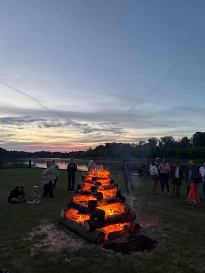 The image depicts an outdoor scene at dusk, featuring a large bonfire constructed from logs. Several people are gathered around the bonfire and in the surrounding grassy area. The bonfire is actively burning, casting a warm glow on the surrounding logs and the faces of some onlookers. In the background, a body of water, likely a river, is visible, bordered by a treeline. The sky above displays a twilight gradient with some cloud formations. The overall atmosphere suggests a communal gathering, possibly a celebration or event, taking place in Carnikava, Latvia, given the context of the location.