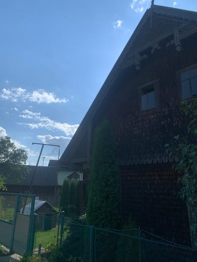 The image depicts a residential property in the city of Yasinia, Ukraine, observed during daytime under a clear blue sky with scattered white cumulus clouds. The primary subject is a multi-story house constructed with dark, overlapping wooden shingles (possibly *dranka* or *gont*) covering its walls and a steep gable roof. The visible facade features two rectangular windows on an upper level and intricate decorative wooden elements along the eaves and at the roof's peak. A house number '7' is affixed to a small, ornamental structure positioned near what appears to be a main entrance, partially obscured by a row of slender, tall evergreen trees.

In the immediate foreground, a dark green metal fence with decorative scrollwork is present, transitioning to a lighter green wire mesh gate on the left. Beyond this gate, a small, light-roofed shed or doghouse is visible near the ground. To the left of the main house, a tall A-frame swing set, constructed from light-colored wooden beams, stands prominently against the sky. A secondary, older wooden building with a dark, sloped roof is partially visible in the background, behind the swing set. The scene contains no discernible human or animal presence, nor any active actions or interactions.