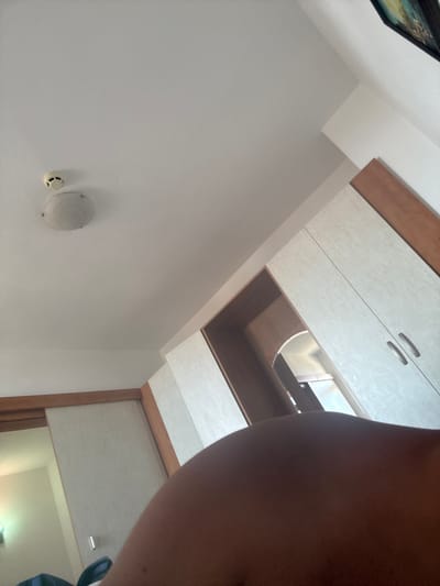 The image displays a bedroom interior, possibly in Sveti Vlas, Bulgaria, viewed from a low angle. In the foreground, a portion of a person's torso is visible, suggesting they are lying down. The room features a ceiling with a smoke detector and a light fixture. To the right, there is a wardrobe with light-colored doors and wooden accents. An open doorway or alcove with a mirrored interior is also present, reflecting some light. The walls are covered in a textured wallpaper. The overall impression is of a domestic interior, possibly a hotel room or apartment.