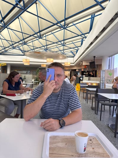 A man with blue eyes and short hair is in the foreground, holding a purple smartphone to his ear. He is wearing a striped t-shirt and a watch on his left wrist. To his right, a white tray holds a takeaway cup with a light brown beverage. The man is seated at a white table, with other tables and chairs visible in the background. The setting appears to be a food court or a casual dining area, with a modern, industrial ceiling structure overhead featuring blue beams and cream-colored panels.
In the middle ground, another man with gray hair is seated at a table, facing away from the camera. Further back, a woman with dark hair is also seated at a table, looking towards the man in the foreground. Behind them, there are various food stalls and signs, suggesting a commercial setting. Visible text includes "FOOD" and "Gracías!" on a sign in the background, and "ONCE" on a yellow sign.
The lighting suggests it is daytime, likely indoors. The overall atmosphere seems casual and relaxed, with people engaged in conversation or enjoying their meals. The image is captured from a slightly elevated perspective, looking down and across the dining area.