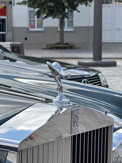A Rolls-Royce car with its signature "Spirit of Ecstasy" hood ornament is parked on a street in Bruges, Belgium. The car's grille and hood are highly polished, reflecting the sky and surrounding buildings. In the background, a building with windows and a tree are visible. The scene is captured in daylight.