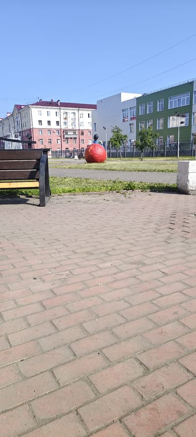 The image depicts a sunny day scene in a public outdoor area, likely a park or a public square in Mezhdurechensk, Russia. In the foreground, a brick-paved walkway dominates the lower portion of the frame, with a dark-colored bench featuring yellow accents positioned to the left. Beyond the walkway, a patch of green grass is visible.
The central focus of the image is a large, bright red, spherical sculpture, possibly representing an apple, with a small figure of a person seated atop it. This sculpture is situated on the grassy area.
In the background, several multi-story buildings are present. To the left, a building with a maroon roof and light-colored facades stands out. Adjacent to it, a modern white building with multiple windows can be seen. Further to the right, a predominantly green building with white window frames and a light-colored facade is visible. A black metal fence separates the grassy area from the buildings in the background, and a few trees are scattered throughout the scene. The sky is a clear, vibrant blue.