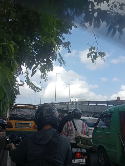 The image depicts a traffic jam scene in Surabaya, East Java. The foreground is dominated by the back of a person wearing a grey hoodie and a black helmet. To their right, another person on a motorcycle wears a distinctive white jacket with colorful striped accents and a green bag slung over their shoulder.

In the middle ground, various vehicles are visible, including a yellow van with stickers on its back, and a green van on the right. The yellow van has text on its rear, including "KURNIA" and "Keluarga Bahagia". Behind these vehicles, a concrete overpass or bridge stretches across the frame, with traffic visible on it. Streetlights and other poles are also present.

The background shows a bright blue sky with scattered white clouds, suggesting it is daytime, likely mid-morning or afternoon. The presence of lush green foliage from a tree in the upper left corner adds to the outdoor setting. The overall atmosphere suggests a busy, perhaps frustrating, moment in urban traffic.