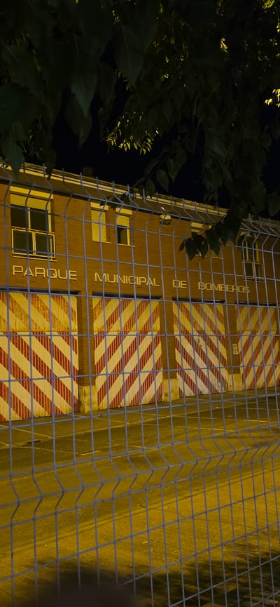 The image shows the side of a brick building at night, with a metal fence in the foreground. The building has several garage doors with diagonal red and yellow stripes, characteristic of fire station entrances. Above the doors, there is text written vertically in large, white, capitalized letters. The text reads "PARQUE MUNICIPAL DE BOMBEROS", which translates to "Municipal Park of Firefighters". The building appears to be a fire station. The time of day is clearly night, indicated by the darkness of the background and the artificial lighting illuminating the building. Some green foliage from a tree is visible on the left side of the image, also lit by the building's lights. The overall scene suggests a functional, likely public building at night in Huesca, Spain.