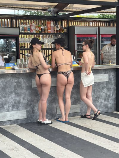 A group of three individuals, two women and one man, are seen at a bar. One woman, wearing a light-colored bikini, stands at the bar, facing away from the camera and adjusting her attire. The second woman, in a leopard-print bikini, is also standing at the bar, looking towards the man. The man, with a beard and wearing a light-colored t-shirt, is behind the bar, interacting with the women.

The bar appears to be outdoors, with a textured, grey stone floor and a tiled floor with alternating grey and white stripes. Behind the bar, shelves are stocked with various bottles of alcohol and glassware. In the background, there are large refrigerators. The scene suggests a casual, social gathering, possibly a bar or a lounge area.

The time of day appears to be daytime, and the weather seems to be warm enough for people to be wearing bikinis. There is no visible text in the image that provides specific information about the location, other than general cues of a bar setting. The overall mood appears relaxed and social.
