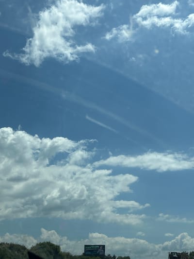 The image displays a wide-angle view of a bright blue sky filled with various types of clouds. Cumulus clouds are prominent, appearing fluffy and white, especially in the lower left portion of the frame. Wispy cirrus clouds are also visible, streaking across the upper part of the sky. In the distance, near the bottom of the image, a billboard with some text and an image can be seen, partially obscured by trees and foliage. The overall impression is a clear day with a mix of cloud formations. There is no visible indication of the city of Lozen, Bulgaria.