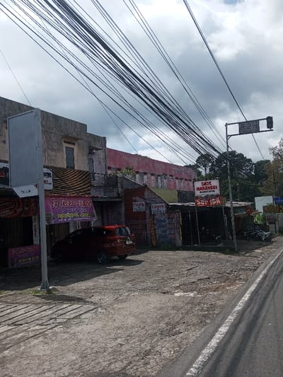 The image depicts an urban street scene, purportedly in the city of Gubbisamamidi, India, though visible signage in the Indonesian language suggests an alternate geographic location. The sky is largely overcast with significant cloud cover. Numerous black overhead wires traverse the upper portion of the frame. The foreground features a worn asphalt road on the right, bordered by a white line, and a dirt/gravel shoulder area extending to the left, where patches of concrete sidewalk are present.
A row of multi-story buildings, exhibiting signs of age and disrepair with faded paint in shades of grey, pink, and red, lines the left side of the street. Some ground-level structures appear to be commercial stalls or shops. A prominent signpost in the left foreground displays "TK LIA" and a large pink banner advertises "ES TELOR JUARA." A dark red hatchback vehicle is parked on the dirt area in front of these establishments.
Further to the right, a white sign identifies a business as "SATE MARANGGI" and "SOP IGA." Below this, faded text includes "Canon" and "WAH BUK." Several motorcycles are partially visible, parked near this food stall. In the far right, a road sign structure with a dark rectangular sign and a traffic signal hangs above the street. Dense green foliage and trees are visible in the background, beyond the buildings on the right. No individuals are visible, and no actions or interactions are depicted within the scene.