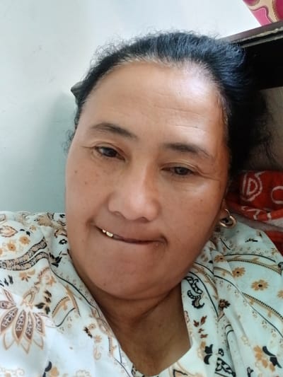 This is a close-up, vertically oriented, selfie-style photo of a woman lying down. She is wearing a light-colored garment with a floral and paisley pattern in shades of white, brown, and black. A portion of another garment, red with a white circular pattern, is visible underneath. The woman has dark hair and is wearing a gold hoop earring in her visible ear. She has a warm, subtle smile. The background appears to be a plain, lightly textured wall, suggesting an indoor setting. The lighting is soft, indicating it could be daytime, possibly indoors near a window. There is no visible text in the image. The overall mood is relaxed and personal.