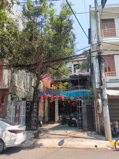 The image captures a street scene in Tuy Hòa, Vietnam, featuring a view of buildings, a tree, power lines, and vehicles. The buildings appear to be mixed residential and commercial, with details like windows, balconies, and architectural styles evident. A large tree with dense foliage dominates the left side of the frame, partially obscuring the buildings behind it. Intricate power lines crisscross the sky, adding to the urban atmosphere. Parked vehicles, including motorcycles and a car, are visible along the street. Signage and decorative elements suggest the presence of a business or establishment, potentially a restaurant or cafe named "SAIGON CA". The overall environment suggests a bustling urban setting with a mix of natural elements and human activity.