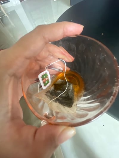 A hand holds a clear glass containing amber-colored liquid and a tea bag. The tea bag has a small, square white tag with a green border and a red design. The glass has a pinkish tint. The background is blurred and appears to be an indoor setting.