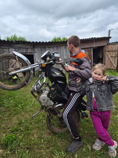 In the city of Kharnavy, Russia, an outdoor scene on an overcast day features a young adult male and a young girl with a black Racer RC50 motorcycle. The male, wearing a grey and orange work jacket, black Adidas track pants with white stripes, and muddy dark sneakers, is seated on the motorcycle, facing left and away from the camera. The motorcycle's front wheel assembly, detached from the forks, is held vertically to the left, displaying a muddy tire and chrome fender. A young girl, approximately 5-8 years old, with light hair, dressed in a dark denim jacket, a colorful t-shirt, bright pink pants, and dirty light-colored sneakers, stands to the right of the male, facing the camera with a playful expression, holding her finger to her chin. The immediate ground is covered with overgrown grass and weeds. In the background, a weathered wooden shed with a dark corrugated roof features a blue circular decal on its wall. One shed door is open, revealing a partially visible individual in a yellow-green top bending inside. Green foliage is visible above the shed, and the sky is uniformly overcast grey.