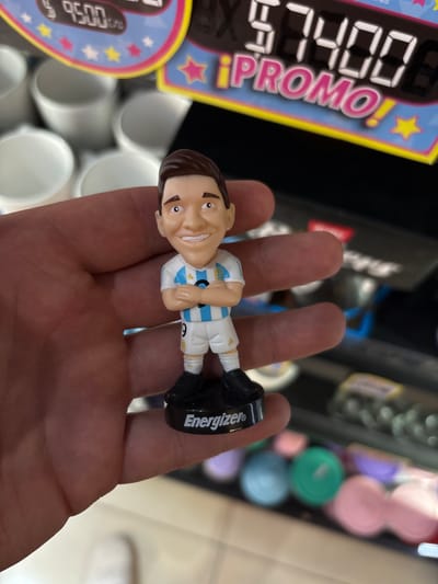 A close-up shot captures a hand holding a small, plastic figurine of a soccer player dressed in an Argentina national team jersey. The figurine stands on a black circular base with the word "Energizer" printed on it in white. The soccer player has a cartoonish, smiling expression. In the background, out of focus, are rows of white cylindrical objects, possibly cups, and a price display board showing "9500" and "57400 ¡PROMO!". This scene appears to be located within a store or a market stall at Aeropuerto Internacional Ezeiza, Argentina.