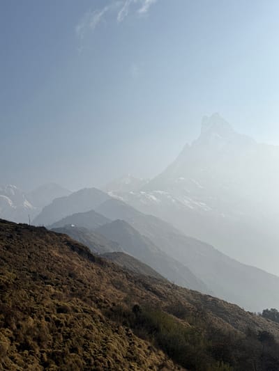 The image shows a mountainous landscape in Lwang Ghalel, Nepal. The sky is a pale blue, fading to white with haze. In the foreground is a steep mountain covered in brown, dry grass and some sparse green vegetation. Behind this, several layers of mountains fade into the distance, their details obscured by mist. The mountains are rocky and rugged in appearance.