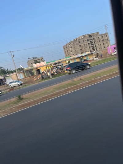 The image depicts a roadside scene in Biashara ward, Kenya, captured from a moving vehicle. The foreground consists of a multi-lane, dark asphalt road with a brown dirt shoulder and a narrow strip of green grass separating it from structures. A dark-colored SUV is in motion on the road, exhibiting motion blur.

On the far side of the road, a row of single-story commercial buildings or market stalls is visible. These structures feature varied facades, including yellow, light green, and light blue. One prominent sign, partially obscured, reads "PREMIUM WHOLESALE...". Several individuals (people) are present on the sidewalk area in front of these establishments, appearing as pedestrians or shopkeepers. Objects consistent with market displays or street vending are situated in front of the shops. A silver or light-colored sedan is parked on the dirt shoulder on the left side of the scene. Utility poles with overhead lines run parallel to the road along the commercial strip.

In the background, a large, multi-story, unfinished concrete building dominates the right side. It features numerous rectangular window openings but lacks finished facades or windowpanes, indicating ongoing construction or disuse. Further to the left in the background, another multi-story building, more finished in appearance, is partially visible. The sky above is clear and light blue. Sparse vegetation is present on the far left and behind some structures.