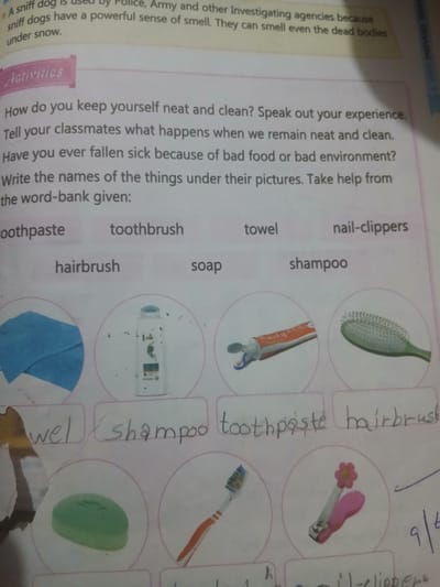 The image depicts a page from a workbook or educational material with text and illustrations related to hygiene and personal care.
The top section contains a paragraph about sniff dogs, followed by a heading "Activities". Underneath, there are questions about personal cleanliness and the consequences of poor hygiene. Below these questions, a word bank is presented with the following words: "toothpaste", "toothbrush", "hairbrush", "soap", "towel", "shampoo", and "nail-clippers".
The lower part of the image features a grid of circular illustrations, each depicting an item from the word bank. These illustrations include:
* A blue towel.
* A bottle of shampoo.
* A tube of toothpaste.
* A green hairbrush.
Below these illustrations are handwritten labels, presumably filled in by a student, matching some of the items to the pictures:
* "wel" (likely "towel")
* "shampoo"
* "toothpaste"
* "hairbrush"
Further illustrations with handwritten labels are partially visible at the bottom, showing a bar of green soap, a toothbrush, and nail clippers.
The image is a photograph of a page, likely from a textbook or worksheet, suggesting an educational context. There is no visible indication of location in Peshawar City Tehsil, Pakistan, or any specific environmental details beyond the white paper of the page itself. The scene is static and consists solely of printed and handwritten text and illustrations. There are no people or actions depicted.