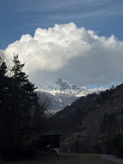 The image depicts a mountainous landscape in Stalden (VS), Switzerland, under a sky with prominent cloud formations. A large, bright white cumulonimbus-type cloud dominates the upper center, partially obscuring the blue sky and casting significant shadows across the lower parts of the scene.

In the mid-ground, a snow-capped mountain peak rises centrally, its upper slopes illuminated by sunlight while its lower flanks are shadowed. Below this peak, a valley with distant structures is faintly visible. To the left, dense, dark coniferous and bare deciduous trees form a shadowed foreground. To the right, a steep, terraced hillside, possibly a vineyard, features sparse vegetation and some scattered small buildings or huts. Faint power lines and poles are visible on the upper right slope.

In the immediate foreground, a traditional wooden chalet-style building with a dark roof and light foundation is partially visible on the left, nestled among shadowed trees. To its right, a patch of grassy ground is present, with a small, white utility vehicle (resembling a golf cart) parked centrally. A narrow dirt or gravel path curves from the right foreground into the scene. No individuals or active interactions are observed. The lighting is high-contrast, suggesting a dynamic weather condition, possibly late winter or early spring.