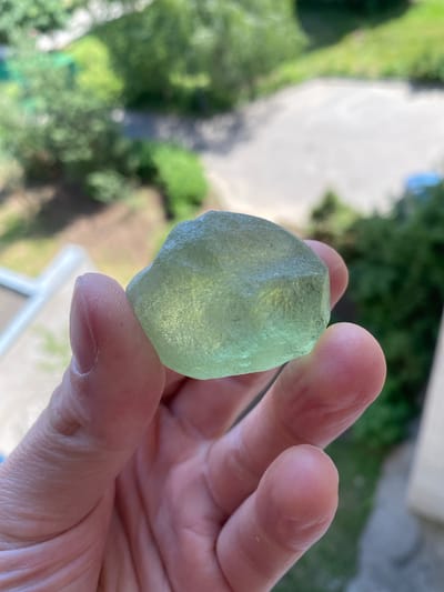 In this close-up shot, a person's hand holds a translucent, pale green, irregularly shaped object, possibly a piece of sea glass or a rough gem. The fingers, with visible lines and texture, gently grip the object, showcasing its frosted surface and slightly angular facets. The background is softly blurred, revealing hints of a natural, outdoor setting. Lush green foliage and the washed-out tones of what appears to be a pathway or ground suggest a daytime scene, likely under natural light. The overall impression is one of casual observation, perhaps of a found natural item. The setting hints at a possibly residential or park-like area, given the greenery and hints of structures. There is no visible text in the image.