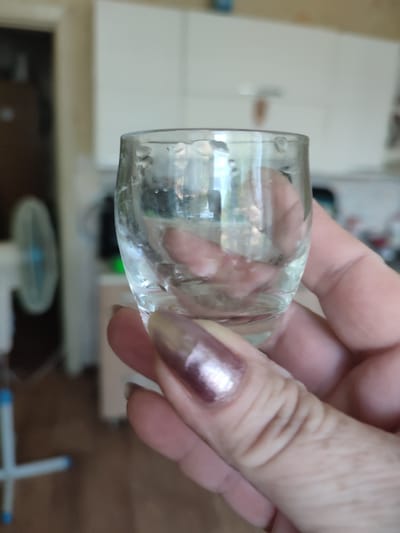 In the foreground, a person's hand holds a small, clear glass containing a small amount of liquid. The glass has droplets of condensation on its exterior. The person's fingernails are painted with a metallic, rosy-purple polish. The background is blurred but appears to be an indoor domestic setting, possibly a kitchen, with white cabinetry and a fan visible on the left. The context of Narowlya, Belarus, is not visually represented in the image.