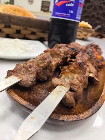 A close-up, eye-level shot focuses on a serving of skewered meat in a wooden bowl on a white tablecloth. The meat, appears to be lamb or beef, is cooked with visible char marks and is presented on metal skewers. To the left of the meat, a white plate holds thinly sliced white onions. In the background, a woven basket contains a golden-brown bread. Behind the basket, a bottle of dark soda is partially visible, with a label that reads "cola." The overall setting suggests a restaurant or home meal in Ivanovo, Russia.