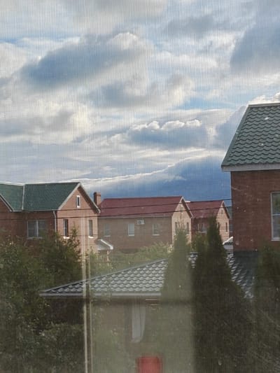 The image captures a view of houses and the sky through a screen, likely from a window. The sky is filled with a dramatic mix of white and dark clouds, suggesting either an overcast day or a storm approaching. The houses are made of brick and have distinctly shaped roofs, with some featuring green shingles and others red. The architecture appears to be suburban. There are trees and bushes visible in the foreground and between the houses, adding to the residential feel of the scene. The screen creates a textured overlay on the entire image, with visible vertical lines. The overall impression is a slightly obscured, but clear, view of a residential neighborhood in a setting with dynamic weather. Based on the provided context, this scene is located in Подстёпки, Russia.