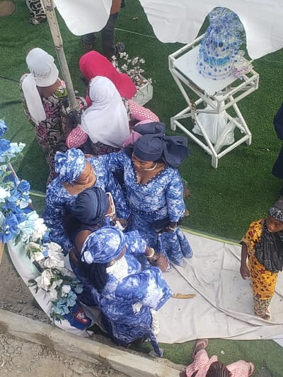 This high-angle image depicts an outdoor social gathering in Ajaokuta, Nigeria. The ground surface consists of green artificial turf and sections of white tarpaulin.
A central cluster of four adult females is prominent, dressed in matching blue and white tie-dye patterned garments and head wraps, engaged in close conversation. One of these individuals holds a mobile phone. To the upper left, three other adult females are partially visible, two wearing white head wraps and patterned dresses (one floral, one pink), and another in a solid red dress. A young child, dressed in a yellow and brown patterned outfit with a dark head covering, stands isolated on the white tarpaulin to the right. A partial view of an individual in pink patterned attire is visible at the bottom edge.
On the right, a white, multi-tiered metal stand supports a wrapped blue and white decorated item, possibly a gift or cake, accompanied by two small decorative bottles. In the lower left quadrant, a white structure is adorned with blue and white artificial flowers. A metal pole, likely part of a canopy structure (partially visible white fabric at the top), extends near the center-left. A concrete ledge forms the immediate foreground.