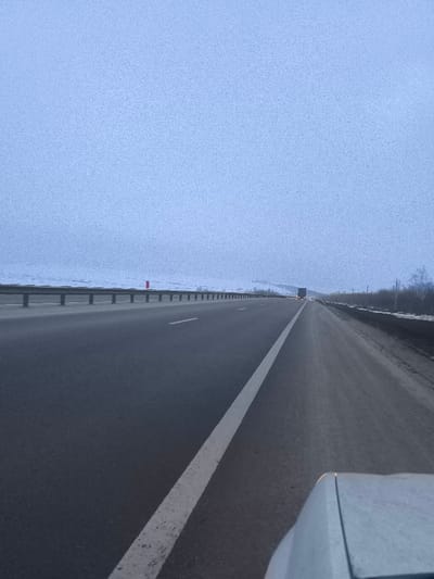 The image depicts a multi-lane asphalt highway scene, viewed from the perspective of a vehicle's dashboard or hood, in what is identified as the vicinity of Pobeda, Russia. The road surface is dark grey, with a prominent solid white line delineating the right edge of the immediate lane, extending into the background. To the left of the road, a metal guardrail runs parallel, separating the highway from snow-covered, undulating terrain that extends to the horizon. A single red, vertical marker is visible near the guardrail in the mid-distance on the left. To the right of the highway, the shoulder appears darker and less snow-covered, transitioning into sparse, bare trees and brush partially visible along the roadside. In the far distance, a semi-truck is visible, traveling in the same direction, with its red taillights illuminated. The sky occupies the upper half of the frame, presenting a uniform, textured light blue-grey, indicating heavy overcast conditions. No people are visible. The overall environment is consistent with a cold, winter day.